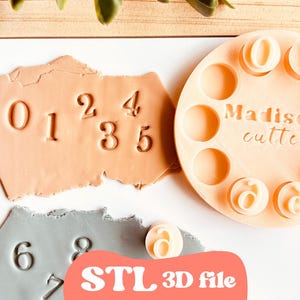 Op de afbeelding: Een lichtoranje cirkelvormige stempelset met de nummers 0-9 en de tekst "Madison cutters". De nummers zijn gestempeld in lichtoranje en grijze klei. De tekst "STL 3D file" staat op een roze banner.