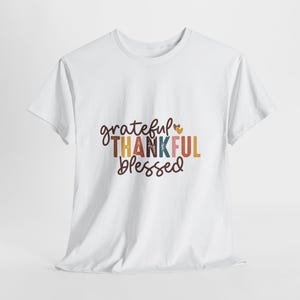 May include: A white short-sleeved t-shirt featuring the text grateful THANKFUL blessed in a multi-coloured palette of brown, orange, blue, and pink. The word grateful and blessed are in a dark brown script font, while THANKFUL is in block letters.