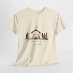 Könnte beinhalten: Ein cremefarbenes T-Shirt mit kurzen Ärmeln und einer aquarellartigen Krippenszene als Grafik. Das Design zeigt einen Stall, Figuren und den Text "For unto us a child is born" sowie "ISAIAH 9:6" mit kleinen dekorativen Blattelementen.