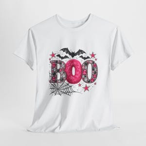 Pode incluir: Uma t-shirt branca de manga curta com uma impressão gráfica da palavra BOO. As letras B e O estão preenchidas com pedras preciosas cor-de-rosa e pretas, enquanto o O central é feito de pelo sintético cor-de-rosa. Morcegos pretos, uma teia de aranha e estrelas cor-de-rosa rodeiam-na.