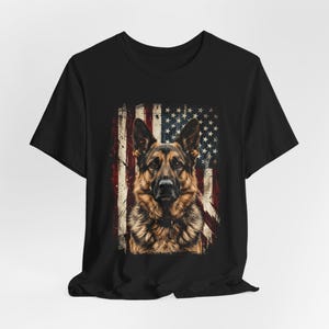 以下が含まれることがあります： 黒いTシャツ。背景に使い古されたアメリカ国旗をあしらったジャーマンシェパード犬のデザイン。犬は茶色と黒色。国旗は赤と白のストライプと、白い星が散りばめられた青い部分があります。愛国的なテーマ。