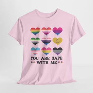 Può includere: Una maglietta rosa chiaro con nove cuori colorati che rappresentano diverse bandiere del Pride LGBTQ+. Sotto i cuori, il testo "YOU ARE SAFE WITH ME" è in nero, con piccole grafiche floreali su entrambi i lati.