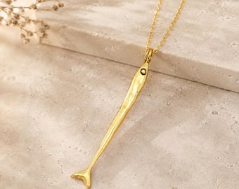 May include: A gold-toned necklace featuring a long, slender fish-shaped pendant with a hammered texture and a small circular eye detail. The pendant hangs from a delicate matching gold-toned chain, creating a sleek and elongated jewelry piece.
