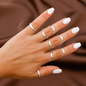 Könnte beinhalten: Eine Hand, die mehrere silberne Ringe zeigt. Die Ringe haben unterschiedliche Designs, darunter schlichte Bänder und offene Kreisformen. Die Nägel sind weiß lackiert, vor einem einfarbigen braunen Hintergrund.