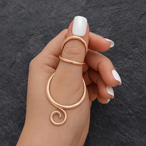 May include: A gold-colored ring set. The set includes a ring for the base of the thumb, a ring for the middle of the thumb, and a large, curved ring with a spiral design. The rings are displayed on a hand with white nail polish.
