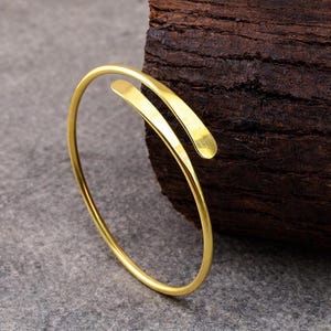 May include: A gold-colored open cuff bracelet. The bracelet has a simple, elegant design with a curved shape and two flat, rectangular ends. The jewelry is displayed on a gray surface with a dark brown wooden background.
