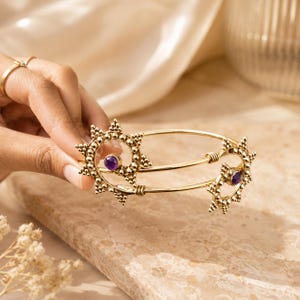 May include: A gold-toned metal bangle bracelet featuring two sun-shaped motifs at the ends. Each sun design is crafted with small beaded details and a central round purple gemstone. The bracelet has a smooth, polished finish and a flexible open design.