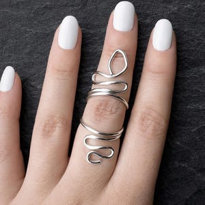 Puede incluir: Un anillo plateado con un diseño único y fluido envuelve un dedo. El anillo tiene una doble banda y un lazo en la parte superior, con un diseño ondulado y abierto. El dedo tiene esmalte de uñas blanco.