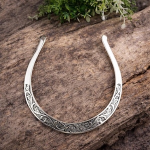 Puede incluir: Un collar plateado con un diseño abierto. El collar presenta un patrón floral grabado a lo largo de la curva inferior. El collar se muestra sobre una superficie de madera con follaje verde en el fondo.