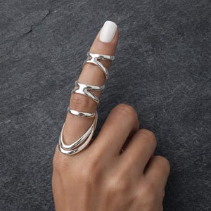 May include: A silver finger ring set. The rings are connected and wrap around the finger. The design includes a curved base ring and several rings that wrap around the finger. The rings are made of shiny silver metal.