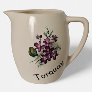 Może przedstawiać: Kremowy ceramiczny dzbanek z uchwytem i kwiatowym wzorem. Wzór przedstawia fioletowe kwiaty, zielone liście i napis "Torquay" czarnym pismem. Dzbanek ma zakrzywiony dzióbek.