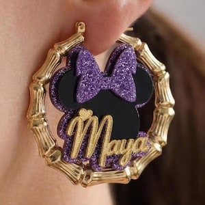 Op de afbeelding: Goudkleurige bamboe oorringen met een zwart silhouet van een muizenkop, een paarse glitterstrik en de naam "Maya" in gouden letters. Een uniek accessoire.