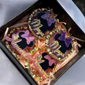 Op de afbeelding: Een set van vier decoratieve oorbellen in een zwarte doos. Elke oorbel heeft een zwart silhouet van een muizenkop met een glitterstrik en een naam in gouden script. De oorbellen zijn omgeven door een gouden bamboe-stijl frame.