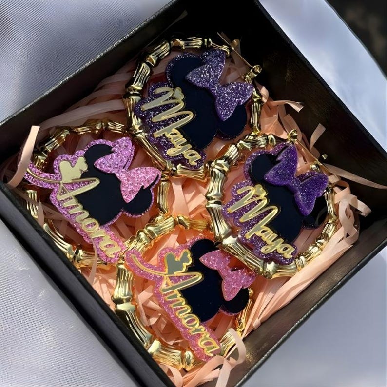 Op de afbeelding: Een set van vier decoratieve oorbellen in een zwarte doos. Elke oorbel heeft een zwart silhouet van een muizenkop met een glitterstrik en een naam in gouden script. De oorbellen zijn omgeven door een gouden bamboe-stijl frame.