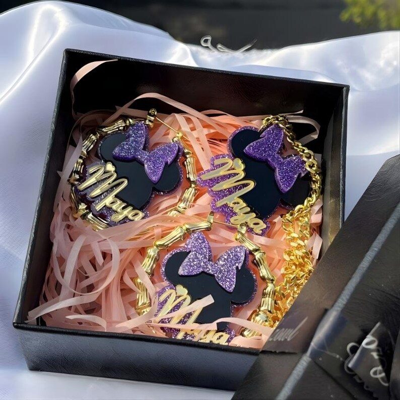 Op de afbeelding: Een zwarte geschenkdoos bevat een sieradenset. De set bevat een ketting en oorbellen, elk met een zwart silhouet met een paarse glitterstrik en de naam "Maya" in gouden letters. De sieraden worden gepresenteerd op roze papier.