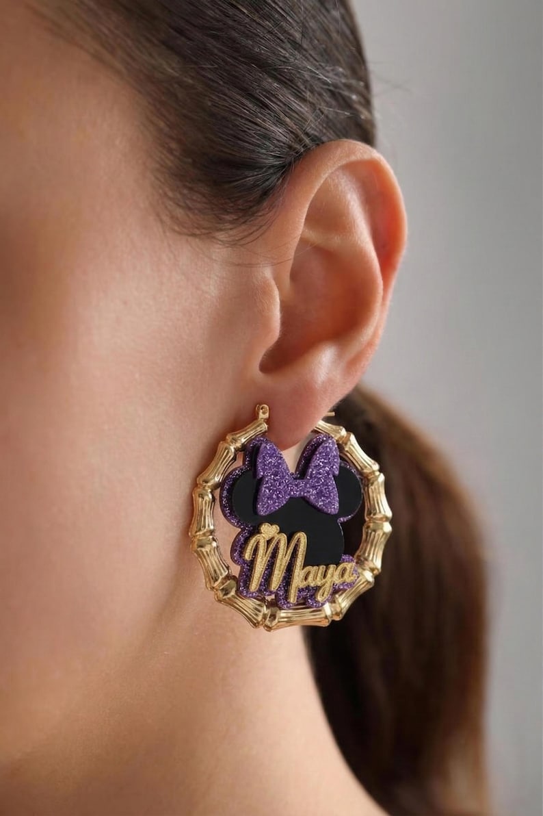 Op de afbeelding: Goudkleurige bamboe oorringen met een zwart silhouet van een muizenkop, een paarse glitterstrik en de naam "Maya" in gouden letters. Een uniek accessoire.