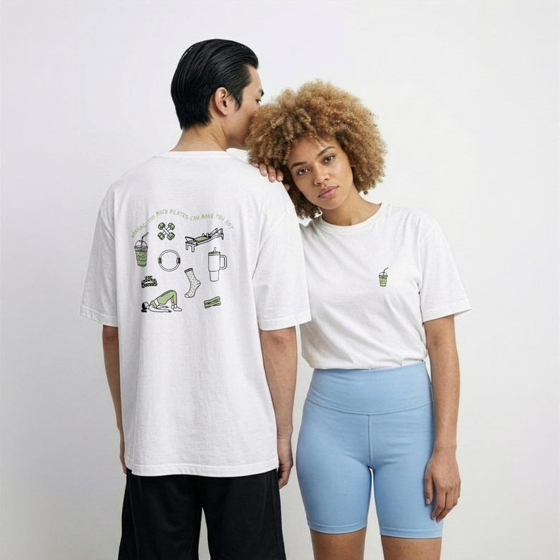 May include: Two people wearing white t-shirts. One shirt has a small green drink graphic, the other has a larger design with fitness-related illustrations and text. The person on the right is wearing light blue shorts. The background is plain white.