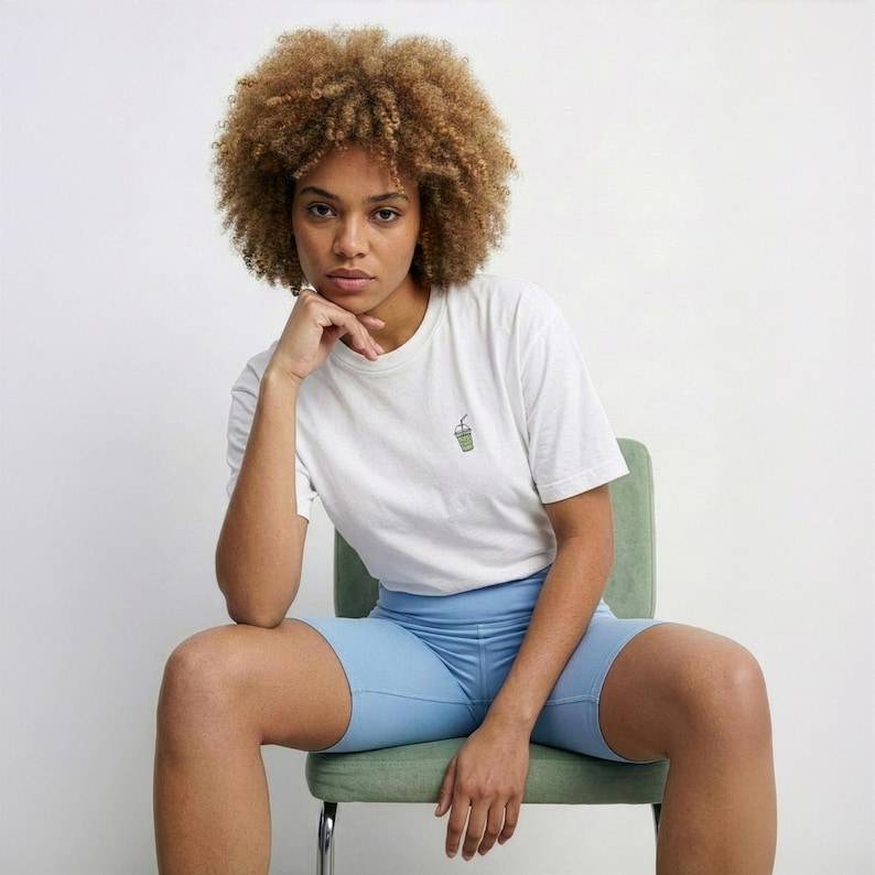 May include: A person wearing a white t-shirt with a small green drink graphic, paired with light blue biker shorts. The person is seated on a green chair against a white background. The t-shirt has short sleeves.