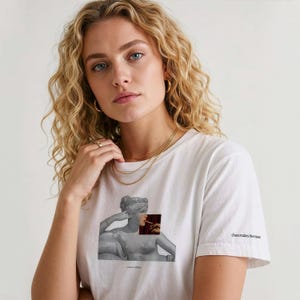 Könnte beinhalten: Weißes T-Shirt mit einer grauen Statuen-Grafik und einem roten und braunen Quadrat. Das Shirt hat den Text "cut & cotton" unten und "chaos makes the muse" auf dem Ärmel. Das Model trägt goldenen Schmuck.