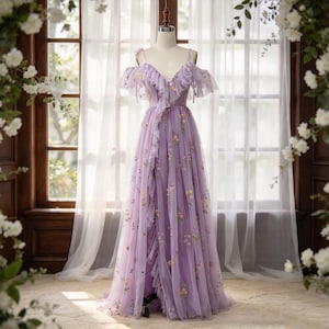May include: A floor-length lavender tulle dress featuring a V-neckline, cold-shoulder sleeves with ruffled trim, and a cascading ruffle down the front. The fabric is adorned with delicate, multi-coloured floral embroidery throughout the bodice and skirt.
