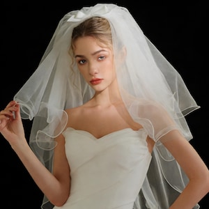 May include: A white bridal veil with a bow at the crown. The veil is made of a sheer, flowing fabric and drapes over the shoulders. The bride is wearing a white strapless dress. The background is black.