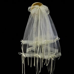May include: A bridal veil with multiple tiers, crafted from sheer white fabric and adorned with delicate lace trim. The veil features ivory satin ribbons and bows, creating a classic and elegant look. The veil is designed to be worn on the head.