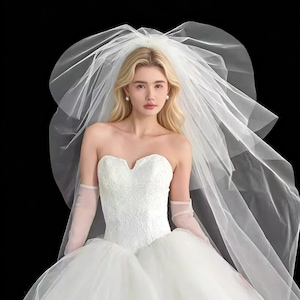 Puede incluir: Un vestido de novia blanco sin tirantes con escote corazón y falda amplia. El vestido presenta intrincados bordados florales en el corpiño. Un velo blanco transparente está drapeado sobre la cabeza y los hombros, creando un look nupcial romántico.