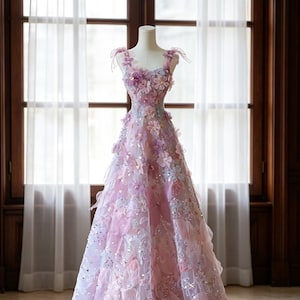 May include: A floor-length sleeveless gown in shades of pink, lavender, and light blue. The bodice and skirt are adorned with three-dimensional fabric flowers, delicate sequins, and small beads. The dress features thin shoulder straps with bows.