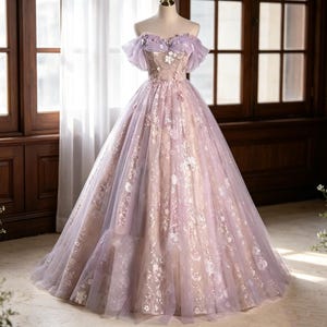 May include: A floor-length ball gown in shades of lavender and soft beige, featuring an off-the-shoulder neckline with ruffled sleeves, a fitted bodice, and a voluminous tulle skirt adorned with intricate floral embroidery and sequin details.