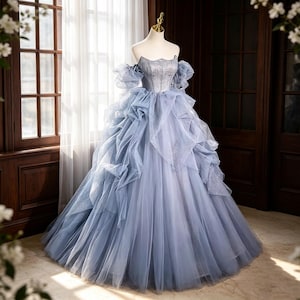 Könnte beinhalten: Ein bodenlanges Ballkleid in Staubblau mit schulterfreiem Ausschnitt, kurzen Puffärmeln und einem strukturierten Oberteil. Der Rock besteht aus mehreren Tüllschichten mit einem dezenten Glitzereffekt und voluminösen, gerüschten Akzenten.