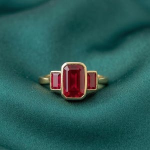 May include: A gold ring with three rectangular, deep red gemstones. The central stone is larger than the two flanking stones. The ring is set against a dark green, textured background.