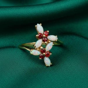 May include: A gold ring featuring a floral design with white opal petals and red gemstone centers. The ring is set against a dark green background, highlighting the contrast of colors and the intricate details of the jewelry.