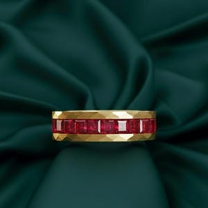 May include: A gold ring with a band of square-cut red gemstones. The ring has a textured, hammered finish on the gold. The gemstones are set in a row around the ring. The background is a dark green fabric.