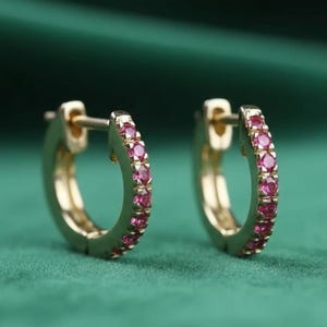 May include: A pair of gold-colored hoop earrings with a row of small, pink gemstones set along the outer curve. The earrings are displayed against a dark green background, highlighting their color and design. The earrings have a classic, elegant style.