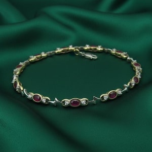 May include: A gold and silver bracelet with oval-shaped red gemstones and small white gemstones. The bracelet is on a dark green satin surface. The bracelet has a clasp closure.