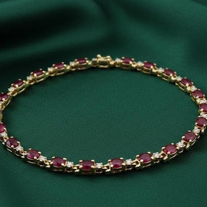 May include: A gold bracelet with oval-cut red gemstones and small white gemstones. The bracelet is set against a dark green background. The gemstones are evenly spaced along the gold chain.