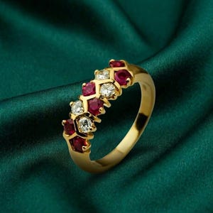 以下が含まれることがあります： 赤と透明な宝石が交互に並んだゴールドリング。 リングのバンドは磨かれたゴールドで、宝石は繰り返しのパターンでセットされています。 リングは、濃い緑色のテクスチャード加工された背景に表示されています。