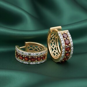 May include: A pair of gold hoop earrings with a row of red gemstones and a row of small white gemstones. The earrings have a geometric design on the inside and are set against a green background.