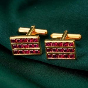 May include: A pair of gold-coloured cufflinks with a rectangular design. Each cufflink features three rows of square, ruby-red gemstones. The cufflinks are set against a dark green, textured background.