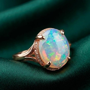 May include: A rose gold ring featuring a large, oval opal gemstone. The opal displays a play of color with flashes of blue, green, and yellow. The band is adorned with small, clear gemstones. The ring is set against a dark green background.