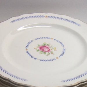 Puede incluir: Un juego de platos de cena de cerámica blanca con un diseño floral azul y dorado. Los platos tienen un borde festoneado y presentan una rosa rosa con hojas verdes en el centro.