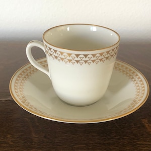 May include: Cream-colored teacup and saucer set with gold geometric trim. The cup has a curved handle and a gold band of triangles around the rim. The saucer also features a gold triangle pattern around the edge.
