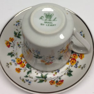 Könnte beinhalten: Weißes Tee-Set mit Blumendesign. Die Tasse ist umgedreht und zeigt den Stempel "Real Brasil by Leart". Die Untertasse und die Tasse weisen ein zartes Muster aus gelben, roten und blauen Blumen mit grünen Blättern auf.