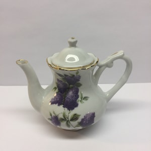 May include: A small white porcelain teapot with a gold trim and a floral design featuring purple flowers and green leaves.