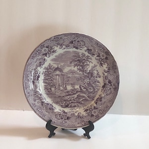 Może przedstawiać: Biało-fioletowy talerz porcelanowy z kwiatowym obramowaniem i centralnym obrazem budynku i drzew. Talerz stoi na czarnym stojaku.