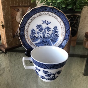 Puede incluir: Un juego de taza y platillo de té azul y blanco con un patrón tradicional de sauce. El platillo tiene un borde dorado.
