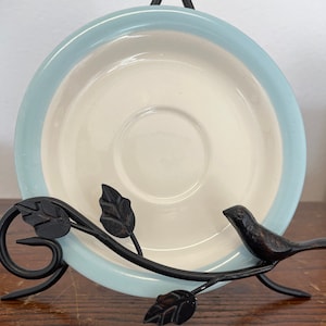 May include: A round ceramic saucer with a cream-colored center and a light blue rim, displayed on a black metal stand featuring a decorative branch motif with sculpted leaves and a small bird perched on the right side of the frame.