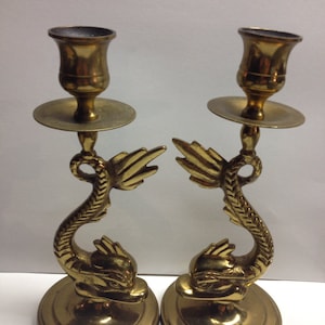 May include: A pair of brass candle holders. Each holder features a dolphin design with a round base, a curved body, and a cup-shaped top. The candle holders are a warm gold colour and have a vintage appearance.