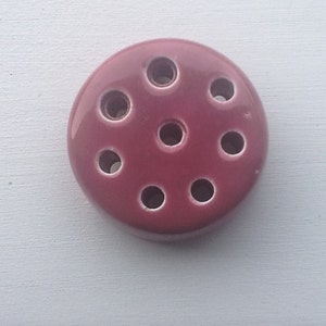 May include: A round, burgundy ceramic flower frog with nine holes for arranging flowers.