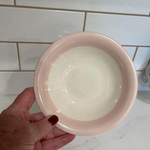 May include: A round ceramic bowl featuring a smooth, cream-colored interior basin and a wide, pale pink rim. The bowl has a simple, circular design with a glossy finish, showcasing a clear contrast between the soft pink border and the light center.